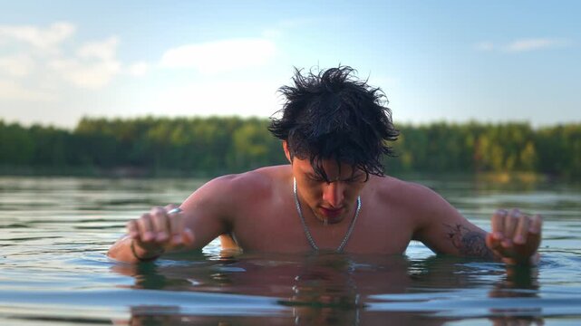 Slow motion low angle view of hispanic fit muscular male coming out of the blue lake water in outdoor nature landscape, wiping his hair, drying while looking around portrait shot.