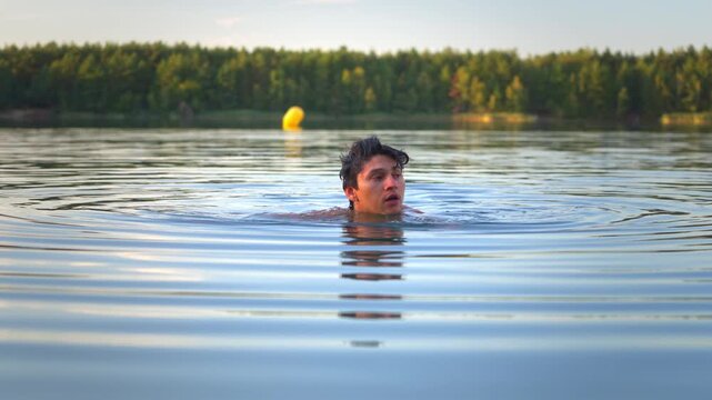 Eye-level medium shot of a young Hispanic man swimming in a serene lake during golden hour, taking a breath and submerging under the clear water surface.