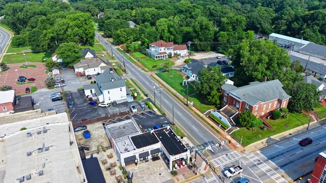 Straight suburban road splits historic brick church from Victorian style homes near Atlanta, Georgia, parked cars line small business lots under dense summer tree canopy. Aerial drone wide shot