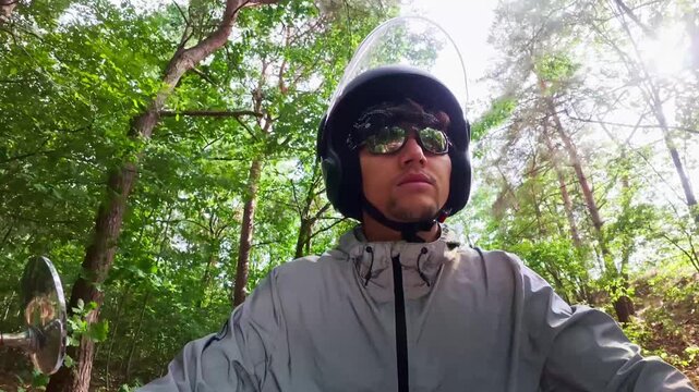 Front-facing medium shot of a stylish young Hispanic man wearing a black open-face helmet, sunglasses, and a windbreaker jacket while riding a moped or scooter along a scenic forest road.