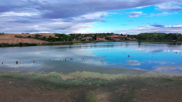 A slower wide camera pan moves across a tranquil lake reflecting bright blue sky and clouds toward a peaceful rural village nestled on the hillside in Als&oacute;mocsol&aacute;d, Baranya County, Hungary.