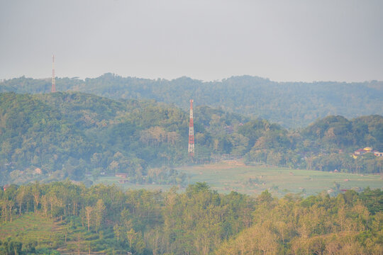 Telecommunication antenna tower standing among lush green hills and rice fields in a hazy countryside landscape under bright sunlight.