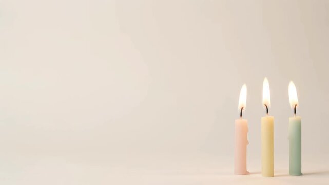 Three colorful thin birthday candles burning with bright flames on plain beige studio background in simple video clip.