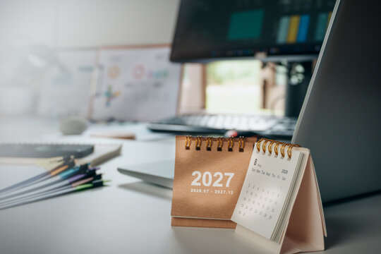 2027 desk calendar on a modern home office workspace with laptop, notebook, stacks of documents and natural window light, representing new year planning, business goals, productivity, future strategy