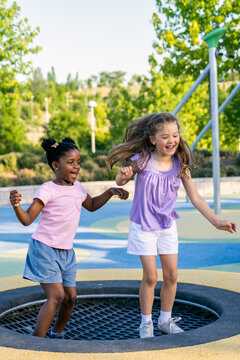 Two multiracial girls laughing and jumping together on a sunny park trampoline, sharing playful energy and childhood friendship