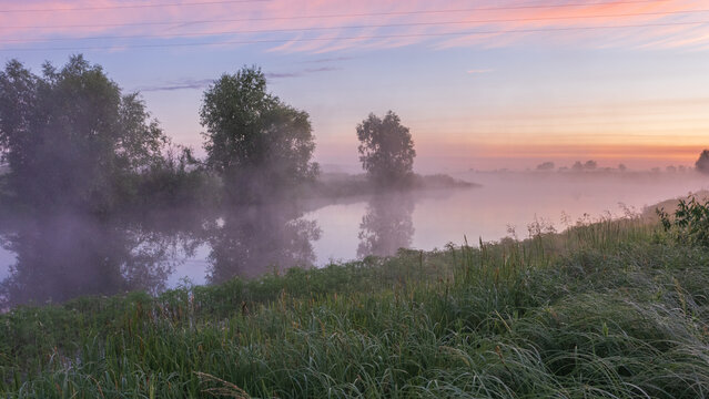 A serene river flows gently under a pastel dawn sky, while thick morning mist wraps around lush green banks and silhouetted trees reflecting softly in the water