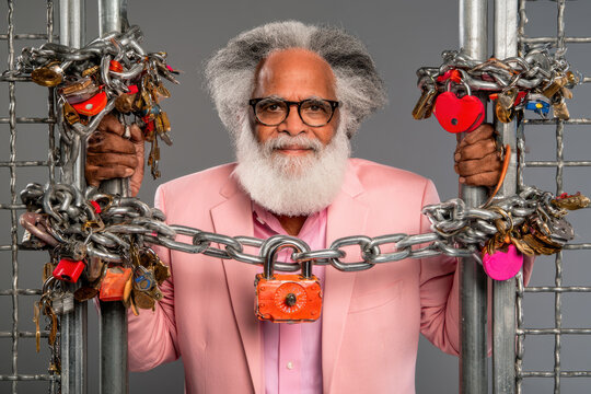 Elderly African American man with white beard wearing pink suit poses between chains and locks, colorful padlocks attached, against a gray background
