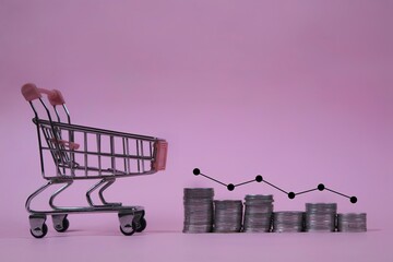 Shopping cart with stacked coins and a declining financial graph, symbolizing consumer spending, purchasing power, inflation, economic downturn, and declining market trends on an pink background. © Ilyas