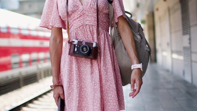 A young woman with a camera and a backpack stands on a railway station platform. Summer trip. Film camera.