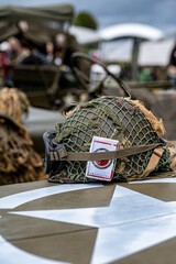 Naklejka premium Sainte-Mere-Eglise, France - June 5, 2026: WWII M1 helmet with net and vintage Lucky Strike cigarette pack tucked under the strap on a military vehicle. D-Day 82nd anniversary commemoration at Camp Ge