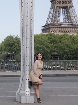 An Asian woman wearing a beige long-sleeved mini dress, sunglasses, and dark boots leans back against a steel pillar bridge, with the Eiffel Tower in the background