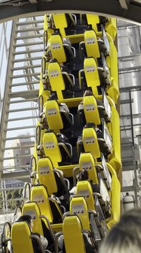 Roller coaster with yellow cars ascends steep track in amusement park, surrounded by modern buildings and trees, showcasing thrilling ride experience in urban setting