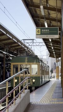 Kamakura, Japan. Green electric train approaches platform at a busy station, with visible signage and passengers waiting along the platform under a clear blue sky