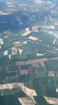 Aerial view of agricultural landscape with patchwork fields and wind turbines, showcasing the gradual transition of terrain and cloud cover over a rural area