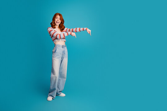 Smiling young woman pointing down with casual striped top and wide jeans on turquoise studio background, cheerful model showing empty space and positive gesture