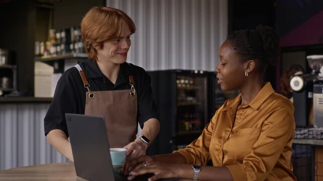 African American young adult woman working on laptop while Caucasian young adult barista serves coffee in cafe. Footage ideal for remote work, digital nomad and hospitality projects