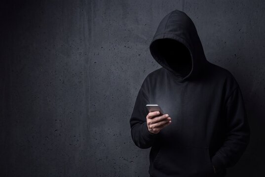 Anonymous hacker in a black hoodie using a smartphone against a dark concrete wall background