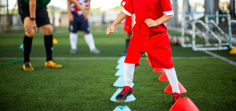 Selective focus to young soccer player in a red kit training on green turf, stepping between agility markers with blurry the supervision of a coach.