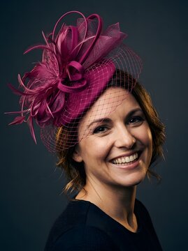 A smiling woman wearing a decorative purple fascinator hat against a dark background.