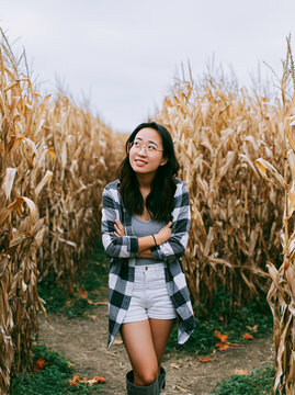 Young asian woman standing and smiling in corn maze on autumn day. Happy girl exploring rural field during fall harvest season. Outdoor adventure and countryside lifestyle concept