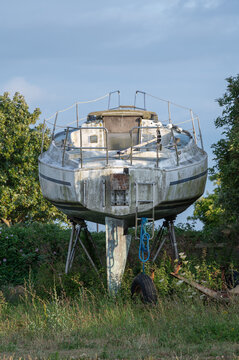 Ein altes, verwittertes Segelboot steht auf St&uuml;tzen an Land in einer &uuml;berwucherten Wiese mit B&auml;umen unter einem bew&ouml;lkten Himmel.