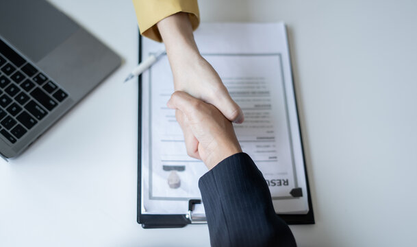 Business partners shaking hands over resume document and laptop at desk for job interview success hiring agreement and partnership concept