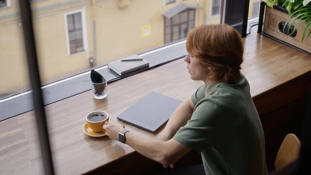 Caucasian young adult man drinking coffee at window counter next to closed laptop, viewed from high angle. Suitable for remote work, freelance lifestyle, and wellness ads