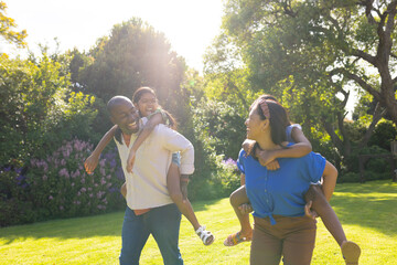 Obraz premium Playing in garden, multiracial parents giving piggyback rides to daughters, smiling and laughing