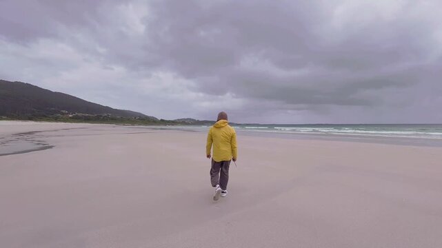 Person in a Yellow Jacket Walking on Carnota Beach in Spain During Overcast Weather
