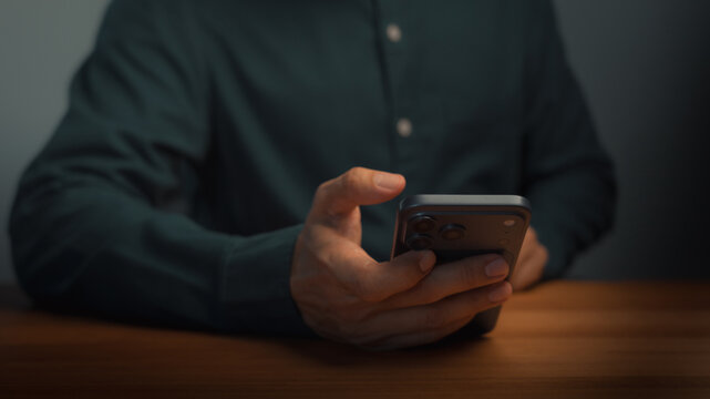 Close up of man hand using smartphone on wooden desk in dark home office. Technology communication concept for online business internet connection and modern mobile network networking.