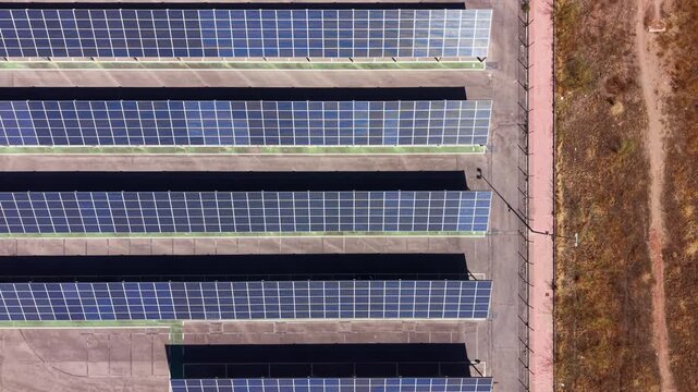 Top-down view of parallel rows of solar panels installed over a parking lot canopy structure, casting shadows on the concrete below beside dry grassland