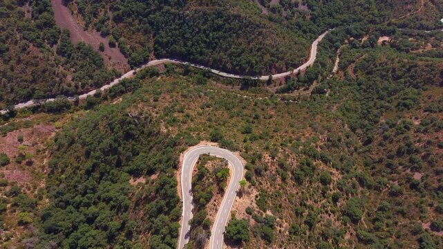 Winding mountain road snaking through dry Mediterranean hillside with dense green shrubland, sharp hairpin bends visible from above on a sunny day