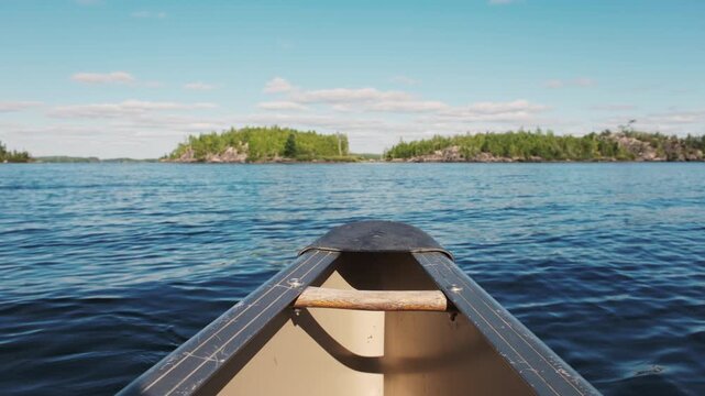 Point-of-view perspective from the bow of a canoe glides across open blue water toward pine-covered islands in the Boundary Waters under a sunny sky.