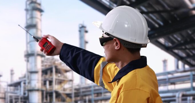Industrial worker in safety helmet and protective uniform using a two-way radio at a refinery, with pipes and processing towers in the background.