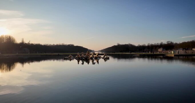 Wide sunset view across Apollo Fountain and the Grand Canal in the Gardens of Versailles, with calm reflections and the historic landscape extending toward the horizon.