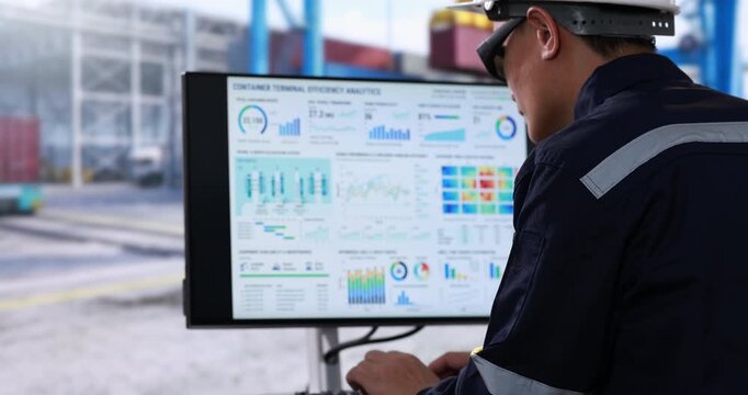 Industrial worker in hard hat and safety glasses reviews data dashboard on a computer monitor at a shipping container terminal.