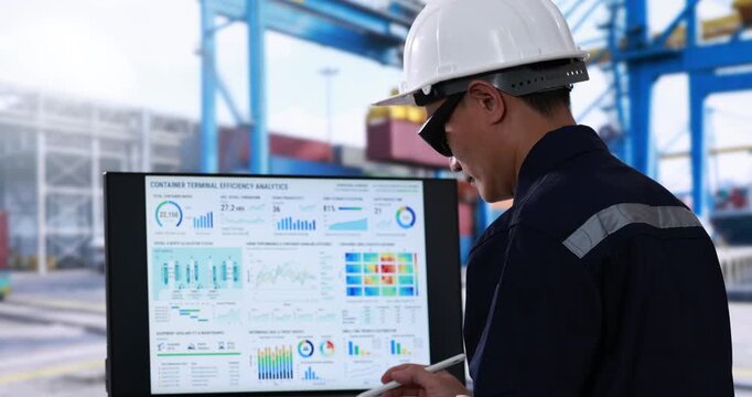 Industrial worker in a hard hat reviews shipping data and colorful charts on a computer screen at a busy container port.