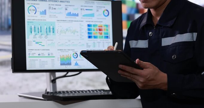 Industrial worker uses a tablet beside a computer monitor displaying colorful production data charts and performance dashboards in a factory office.
