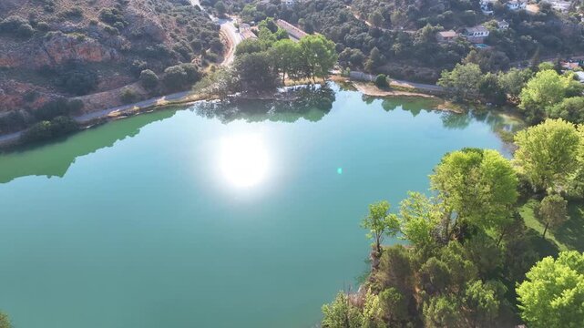 Flight reveals starting blue green lagoon surrounded by lime coloured trees as the hot white globe of sun reflects on the water's surface, Lagunas de Ruidera Castilla La Mancha Spain.