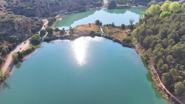 Aerial retreats across two linked Ruidera lagoons and captures a starting column of early morning sun glint from the base of waterfalls. Castilla La Mancha Spain