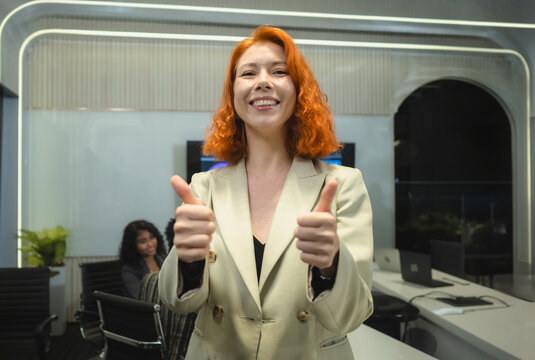 Confident Young Businesswoman . smile ginger-haired female professional blazer show thumbs-up in modern high-tech office representing corporate success positive results professional approval.