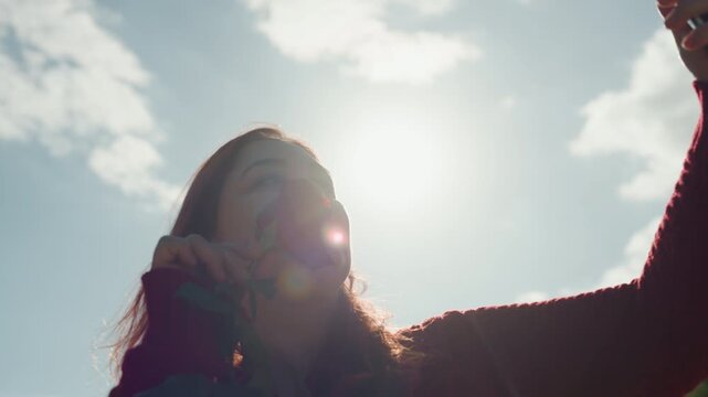 Close up of woman holding red rose and taking selfies with her smartphone outdoors under bright sunlight. Woman wears long sleeved sweater capturing moments against backdrop of trees and sky