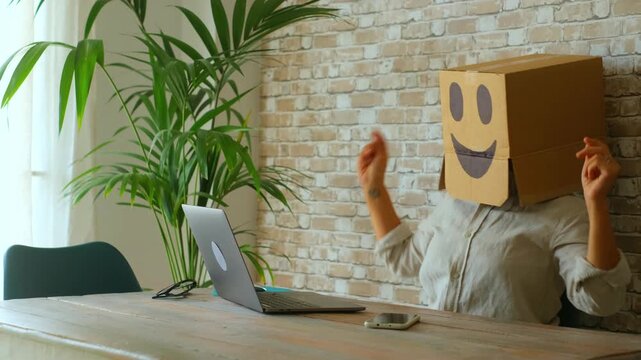 Jubilant businesswoman wearing a cardboard box with a smiley face celebrating success by dancing in her chair after finishing her work on the laptop, expressing happiness and job satisfaction