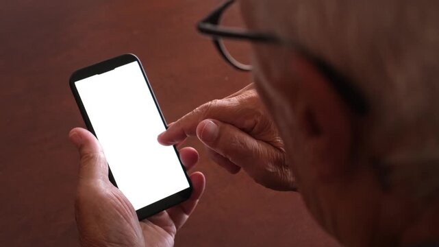 Over the shoulder view of an old man with wrinkled hands scrolling and tapping on a smartphone. The mobile phone has a white screen for easy compositing, ideal for advertising or mockups