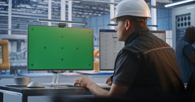 Male Engineer in Hardhat Types at Keyboard Before Two Glowing Chroma Key Green Screen Monitors in Smart Factory. Ideal For Compositing Customized Industrial Software or Telemetry Interfaces.