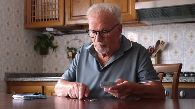 Worried senior man with eyeglasses sitting at the kitchen table counting his last euro banknotes and coins, representing concepts like poverty, financial crisis, and economic hardship