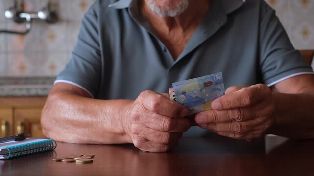 Senior retired man sitting at a kitchen table carefully counting his last savings, holding euro banknotes and separating coins, highlighting the challenges of financial insecurity and poverty
