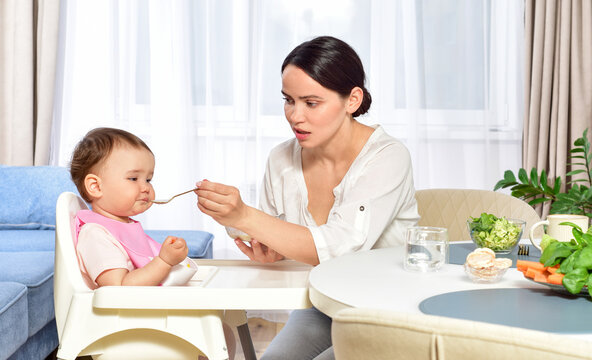 Mother feeding baby with a spoon. Spoon-feed. Happy young mother with a baby in the kitchen interior. Fresh vegetables and fruits.