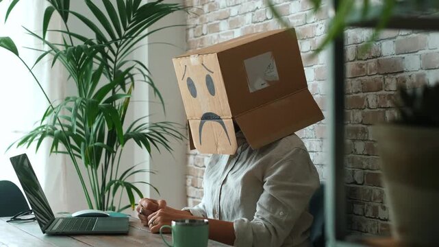 Sad employee wearing a carton box with a drawn sad face on their head, feeling frustrated and stressed while working on a laptop computer in a modern office environment