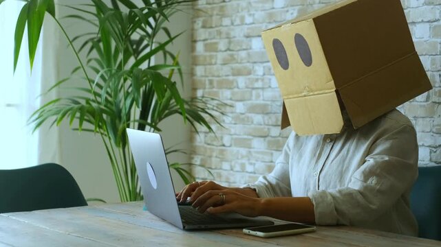 Unrecognizable female employee with a cardboard box on her head typing on a laptop, representing the concepts of online privacy, digital identity, anonymity, and corporate conformity